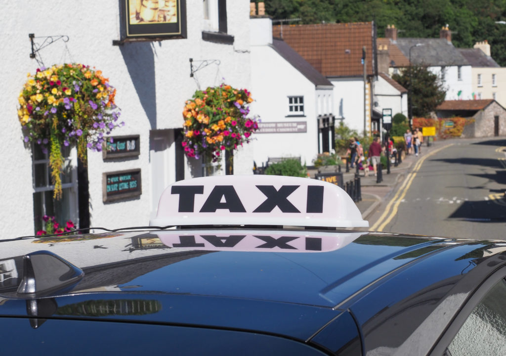 Taxi sign on a vehicle parked in a village street lined with hanging flower baskets, shops and residential buildings.