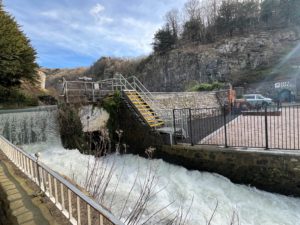 Fast flowing water over a weir with a car park in the background